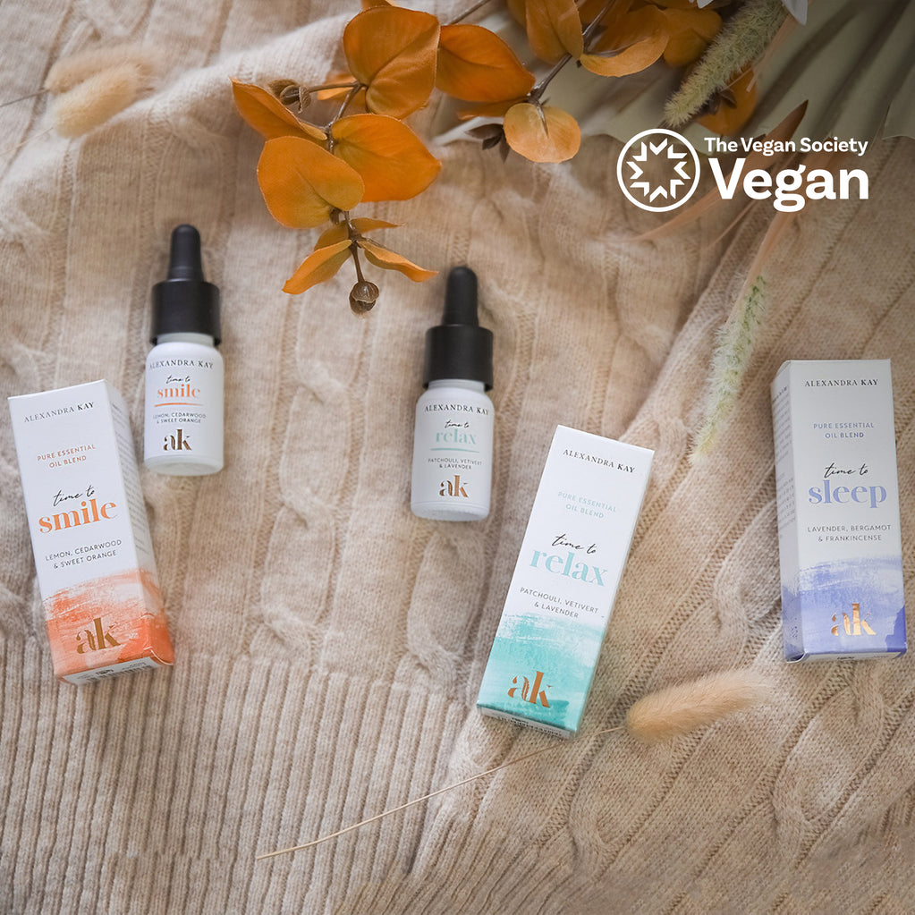 Tres frascos y cajas Alexandra Kay (Time to Smile, Relax, Sleep) sobre un suéter de punto beige con hojas naranjas y el logo de The Vegan Society.