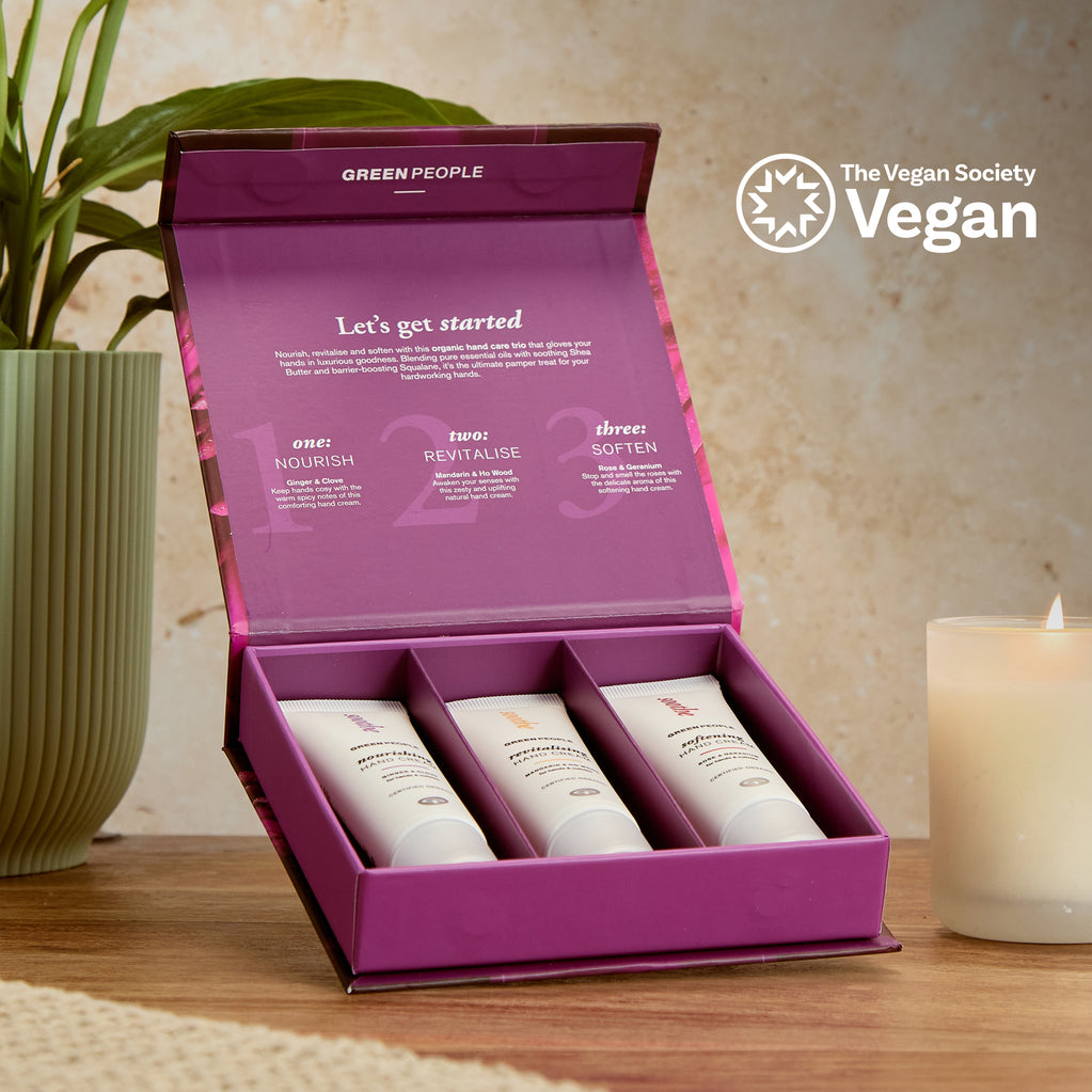 A purple box containing three Green People hand creams sits on a wooden table next to a white candle and green plant. Text explains the trio’s Nourish, Revitalise, and Soften steps.