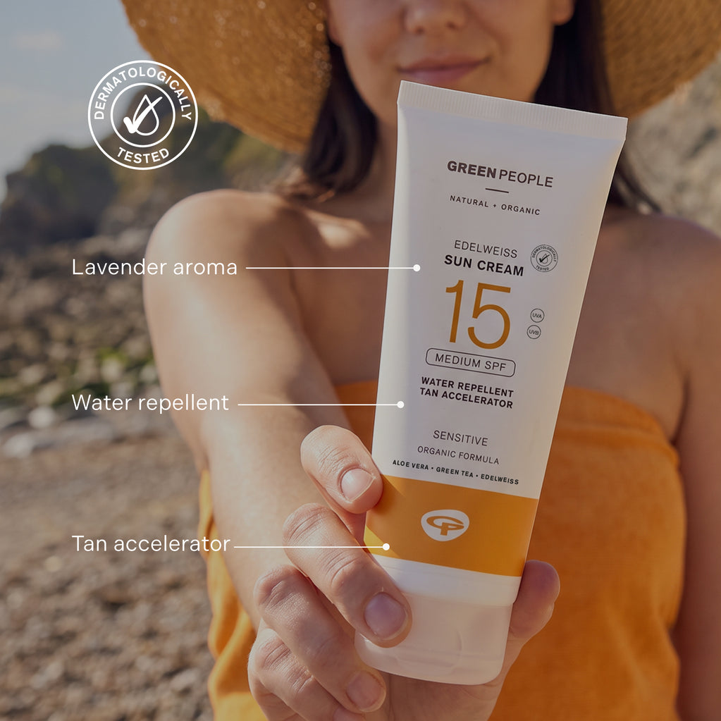 A hand holds a tube of Edelweiss Sun Cream with Tan Accelerator SPF15 on a sunny beach in front of a woman in a straw hat.