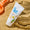 Tubo blanco de crema solar SPF50 en la arena junto a una pala de plástico naranja, con la esquina inferior izquierda recortada.