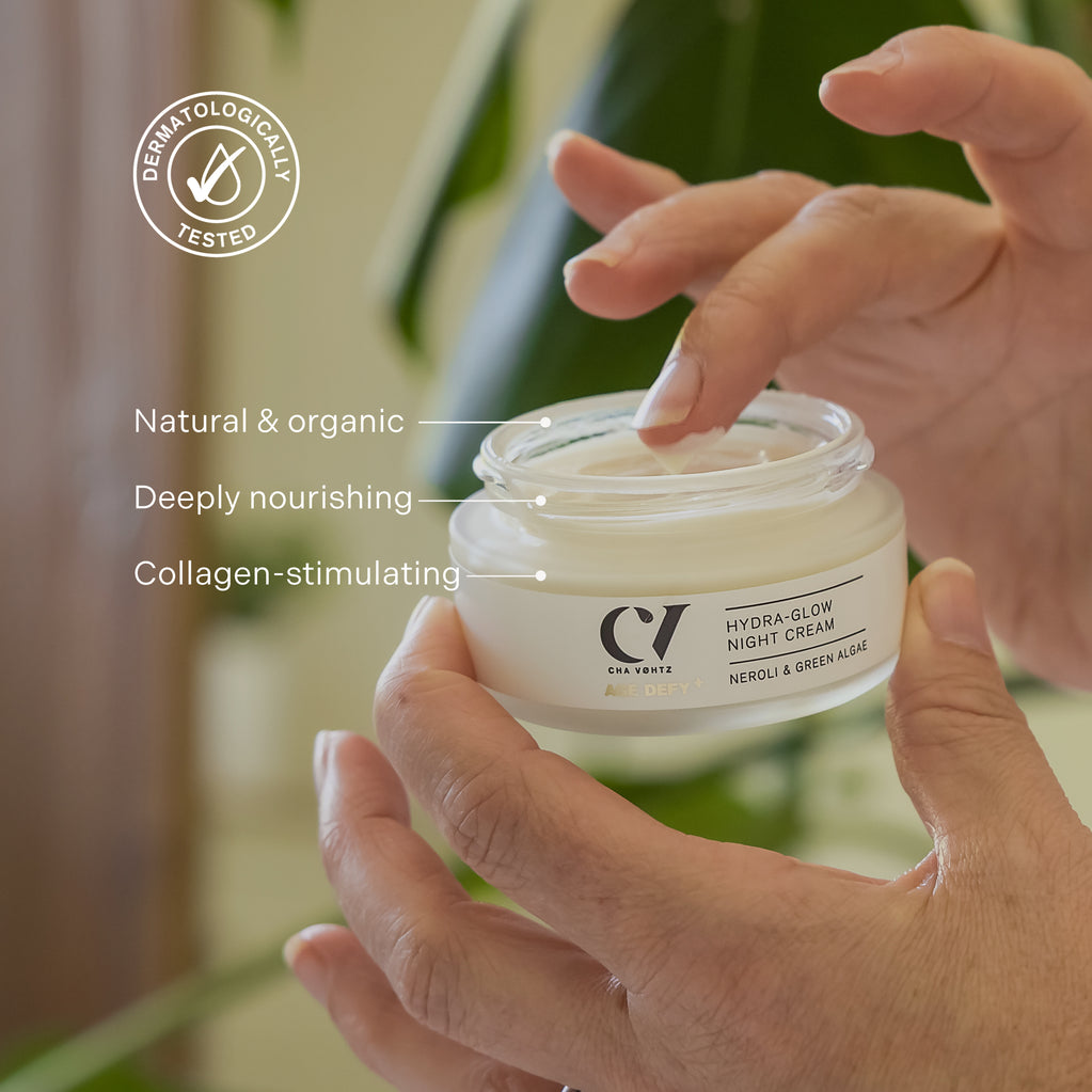 Hands hold an open jar of Age Defy+ Hydra-Glow Night Cream, dipping a finger into the rich white moisturiser.